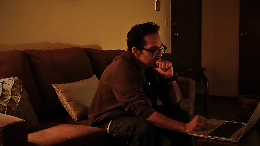 The same man in profile, hand resting near his chin, looking at a laptop screen on the same couch.