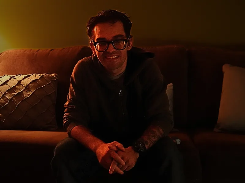 A man with dark hair and glasses sits on a couch, hands clasped, smiling toward the camera in warm, dim lamplight.