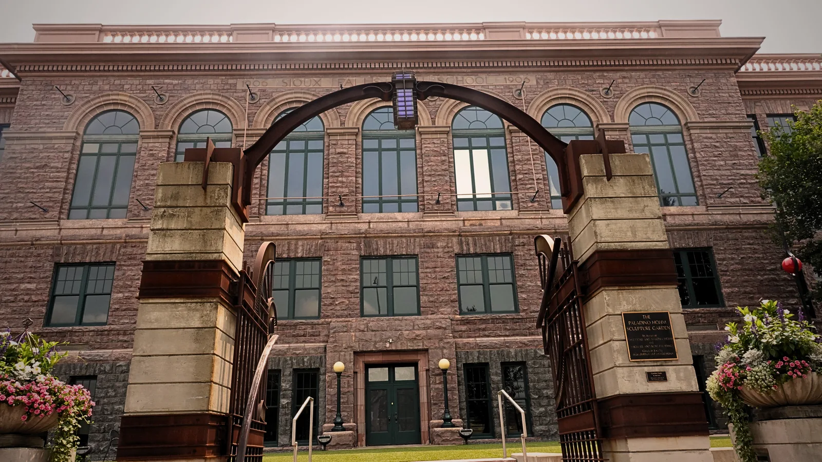 A Sioux quartzite school building in downtown Sioux Falls, dated 1900 and 1906 in the stone above its arched windows, seen through the steel arch of a sculpture garden.