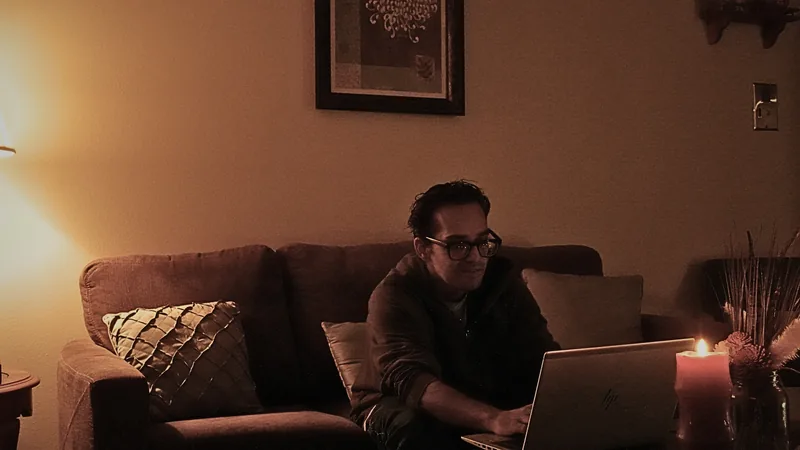A dim, warm-lit living room: a couch, a bright table lamp, a lit candle, and a laptop open on a low table, with a man seated at the laptop.